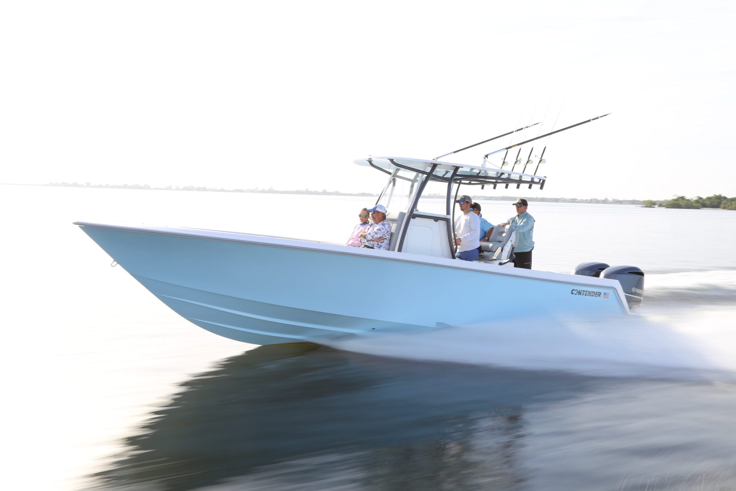 A light blue speedboat with four passengers cruises on calm water, two men seated at the front and two men standing at the steering area, with a distant shoreline visible.