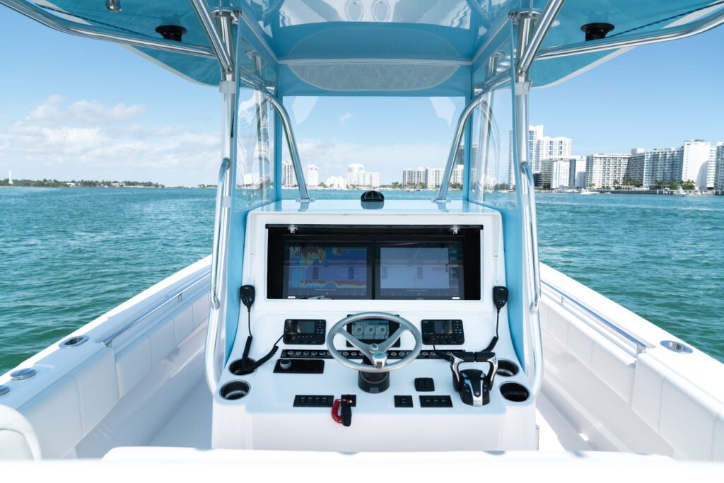 A view of the helm of a modern boat with various controls and displays, navigating through calm waters towards a cityscape of high-rise buildings in the background.