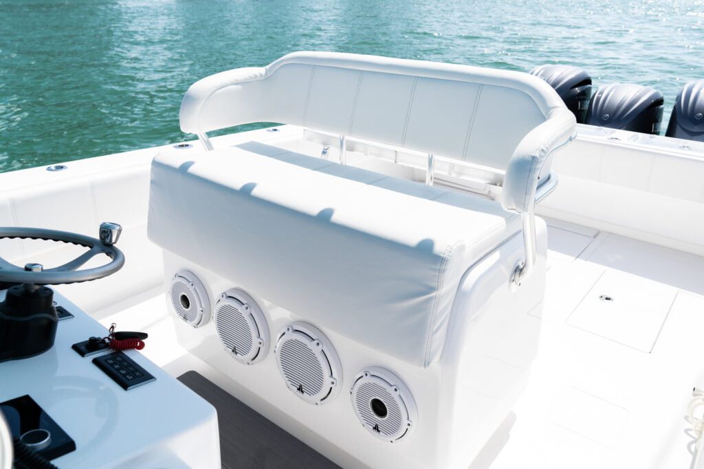 A white boat seat with an upholstered cushion is positioned in front of four circular speakers on a white boat deck, with a ship's wheel visible on the left side and water in the background.