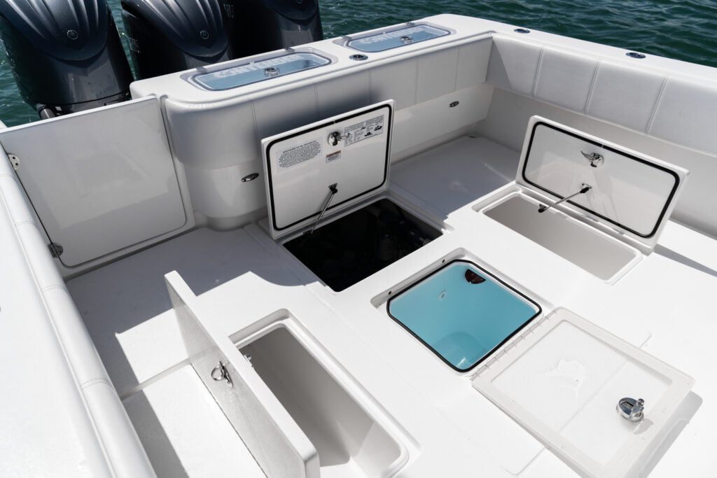 A white boat's deck with multiple opened storage compartments and a view of three outboard engines in the background. The compartments show various storage spaces and access panels.