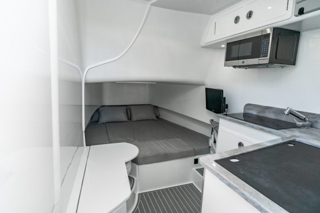 Compact living space with a small bed, kitchenette featuring a microwave, a sink, cupboards, and a small TV. The interiors are modern with white walls and grey accents.