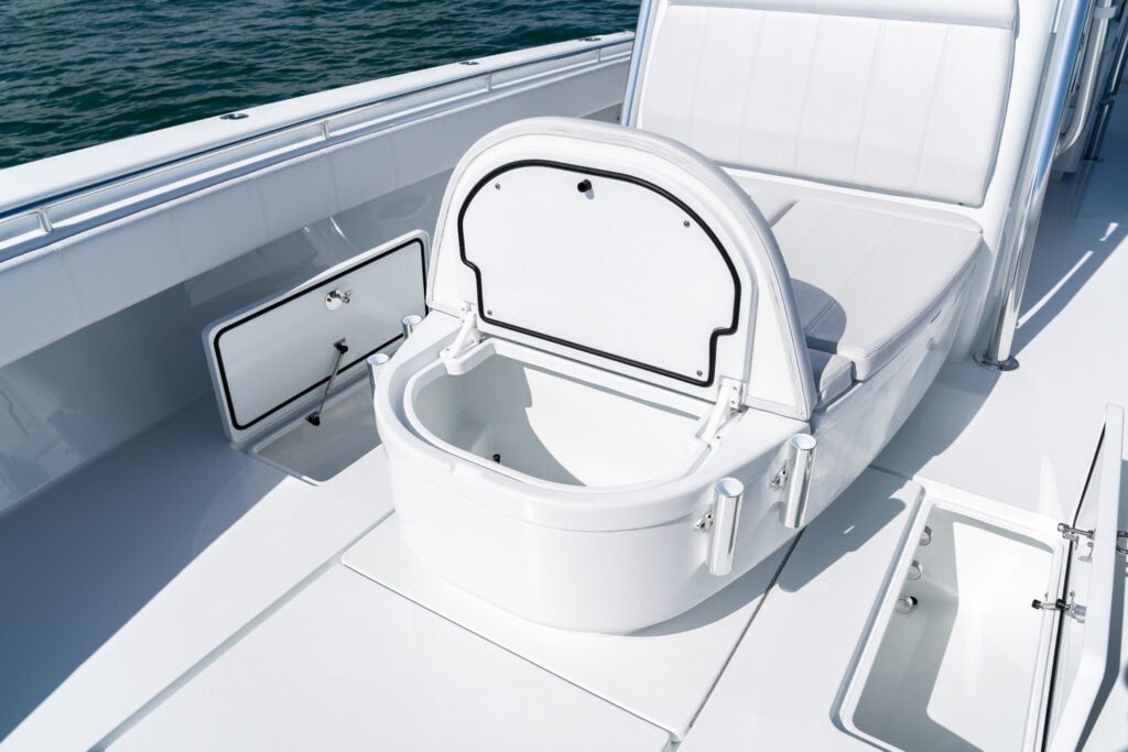 A clean, white open storage compartment on a boat, revealing an empty interior. It is surrounded by additional storage compartments on the deck. The sea is visible in the background.