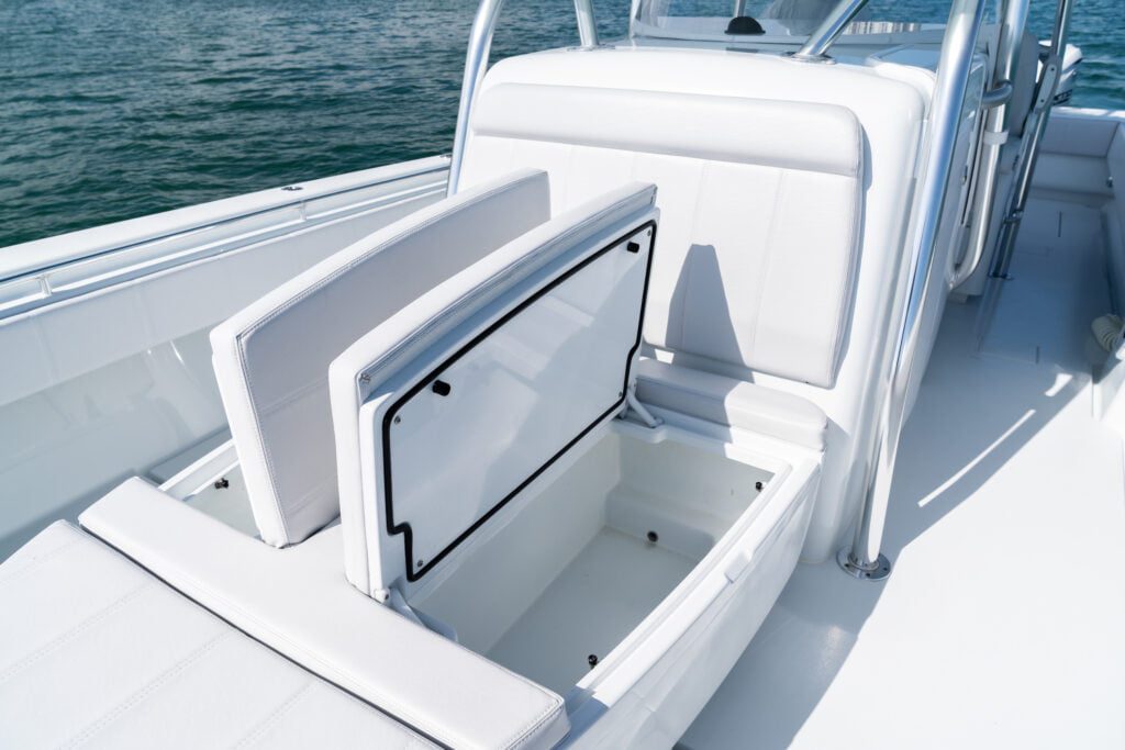 A storage compartment is open on a white boat with seating and railings, positioned on calm water.