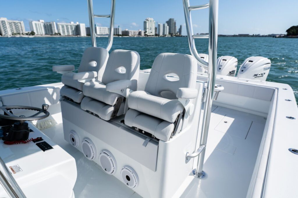 A luxury boat with white cushioned seats overlooks a city skyline. The boat is equipped with multiple outboard motors in the rear, floating on calm blue water under a clear sky.
