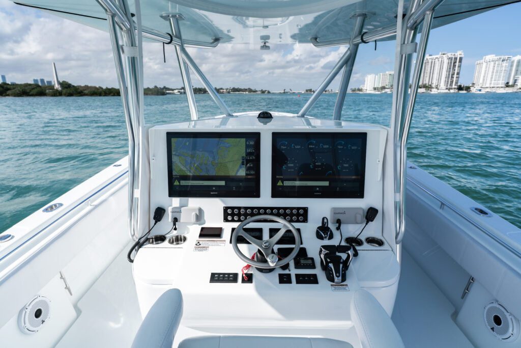 The image shows the control console of a boat with two large navigation screens, multiple controls, and a steering wheel. The boat is on a body of water with buildings visible in the background.