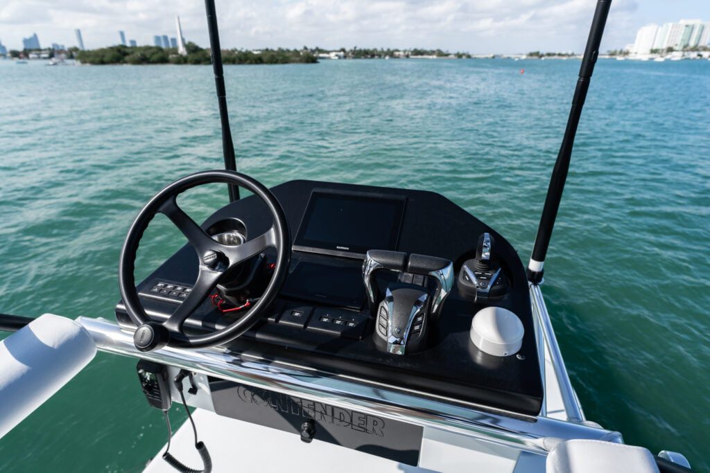 A boat cockpit with steering wheel, electronic controls, and various navigation instruments, offering a view of the water and distant shoreline under a partly cloudy sky.