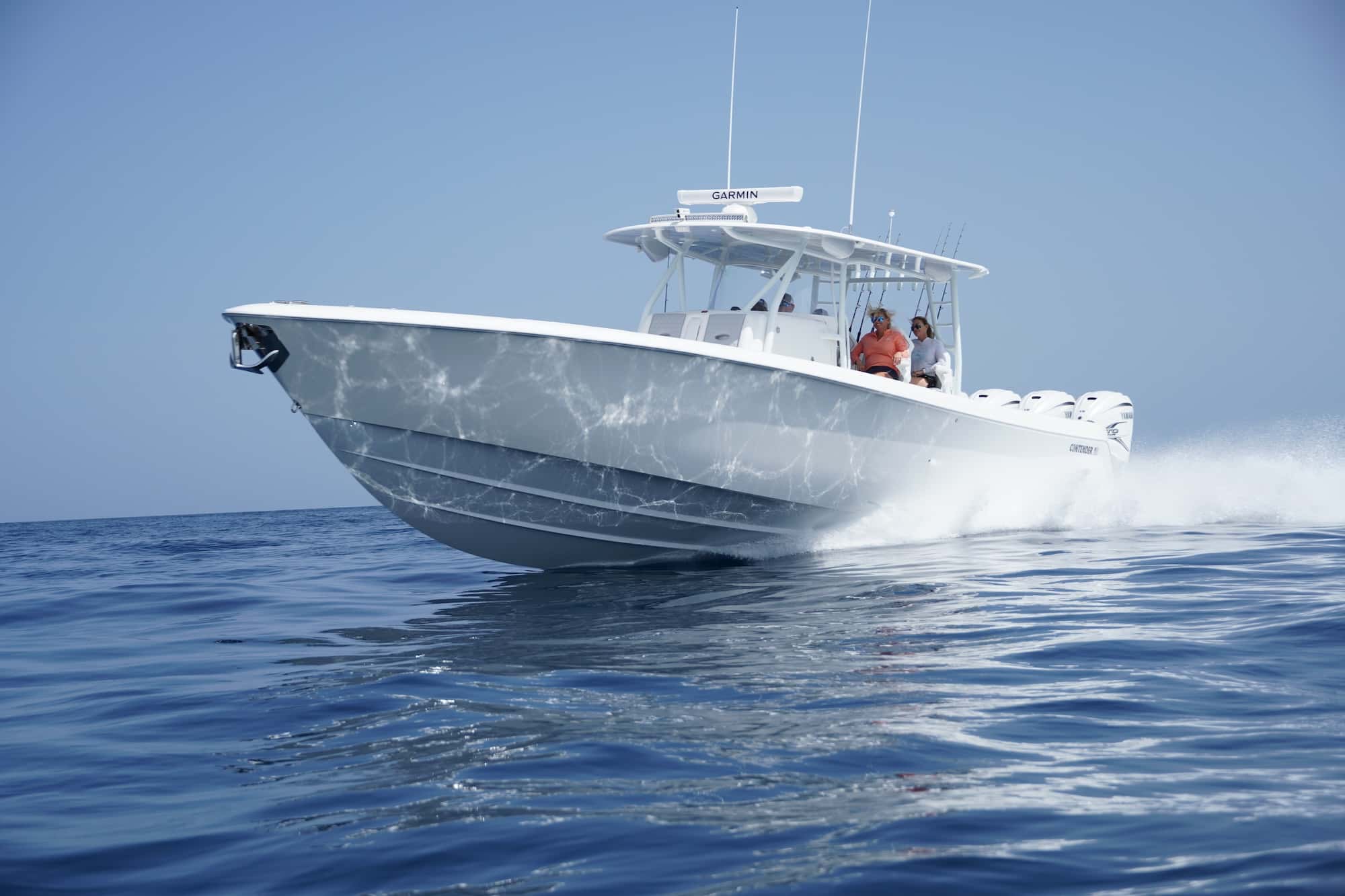 A white motorboat speeds across calm ocean water under a clear sky, with a few people visible onboard.