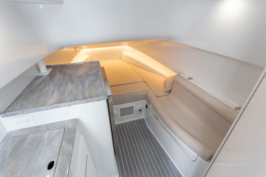 Interior of a boat cabin featuring a small kitchenette with a countertop on the left, a compact seating area on the right, and a sleeping berth at the far end illuminated by soft lighting.