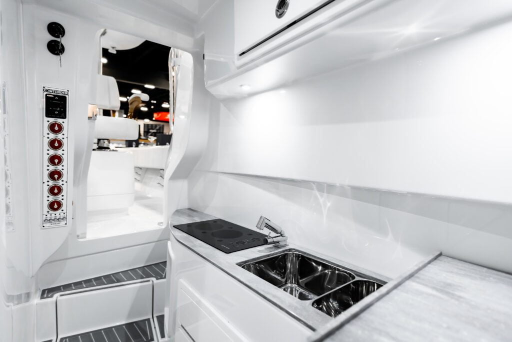 A modern, white RV kitchen area with a two-burner cooktop, sink, countertop, and storage cabinets. Doorway in background leads to living area.