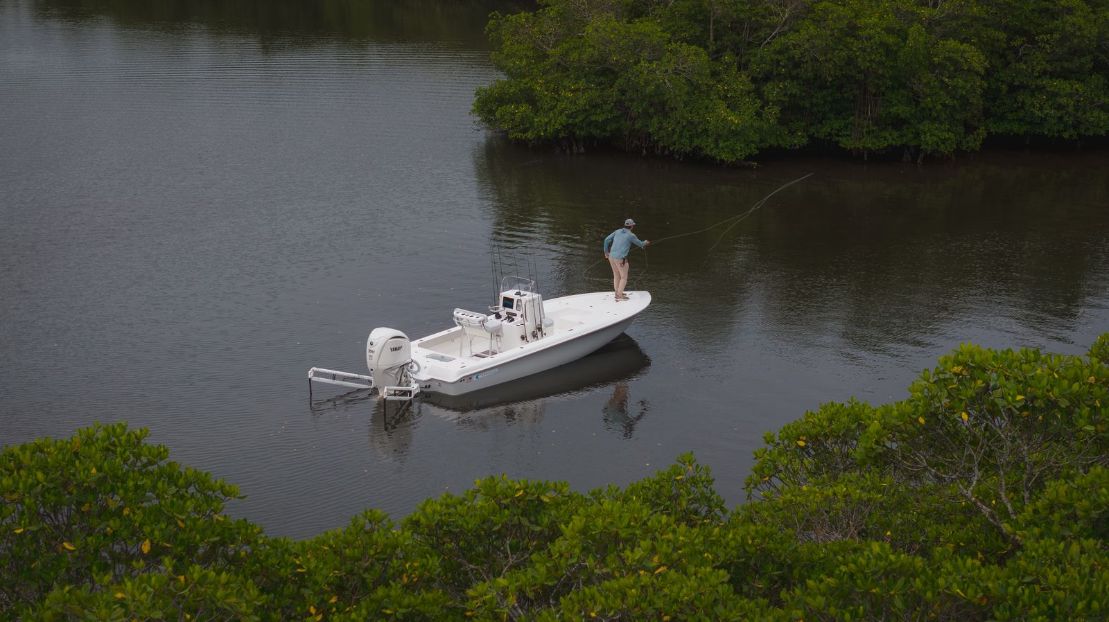 A person stands on a white boat while fly fishing in calm waters near a dense area of mangrove trees.