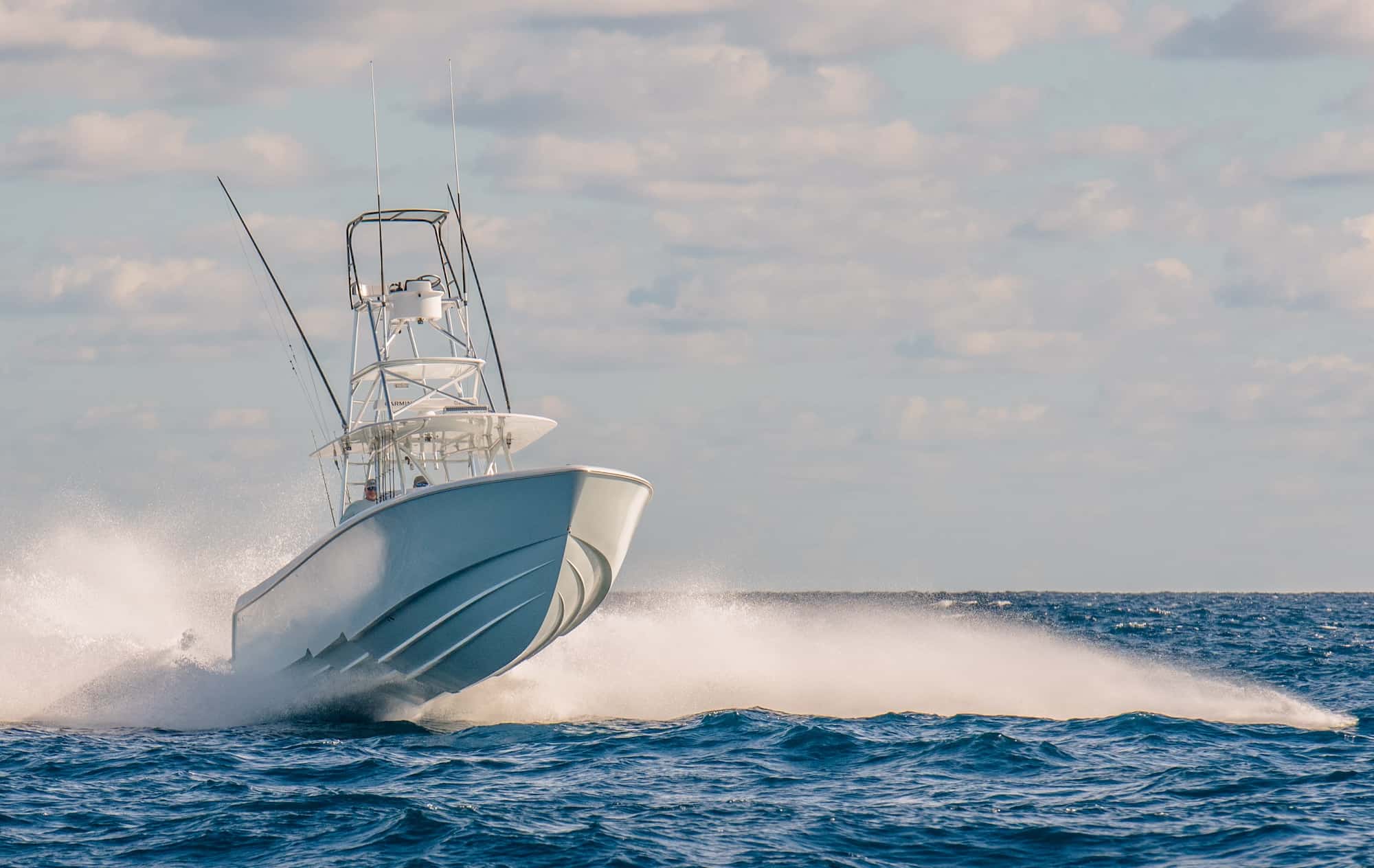 A white motorboat speeds across the ocean, creating a large wake as it moves through the water under a partly cloudy sky.