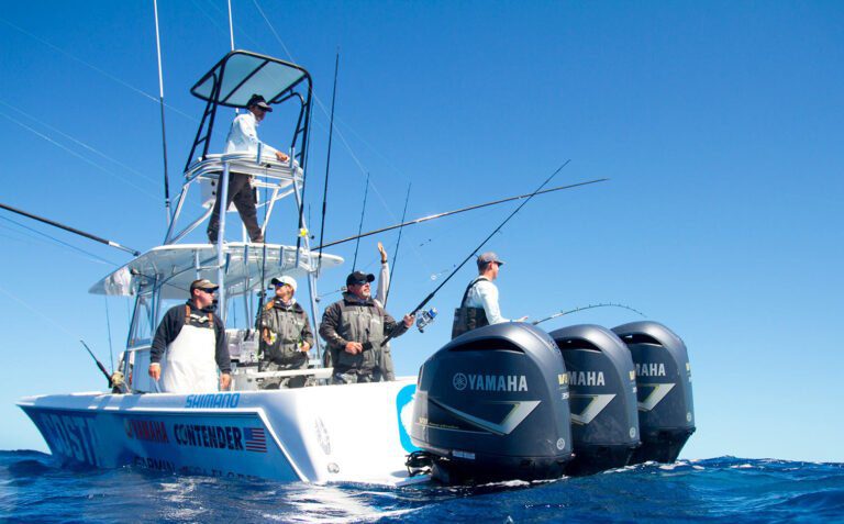 A group of people fishing on a motorboat with multiple rods in the water. The boat has Yamaha engines and various sponsor logos. The water is calm and the sky clear.