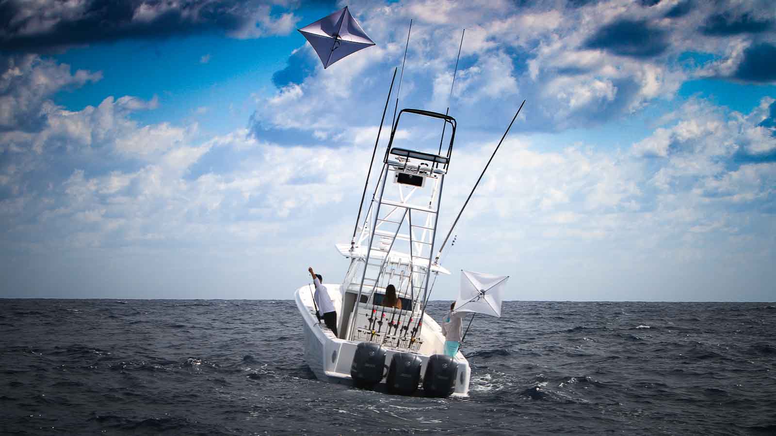A white fishing boat with three outboard motors lists to one side in open water under a cloudy sky, with two kites flying above.