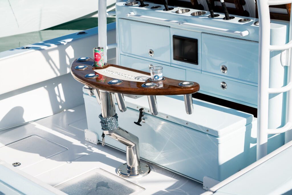 A modern boat's cockpit area featuring a central fishing station with rod holders, a wooden table, and drink holder. A soda can and a drink cup are on the table.