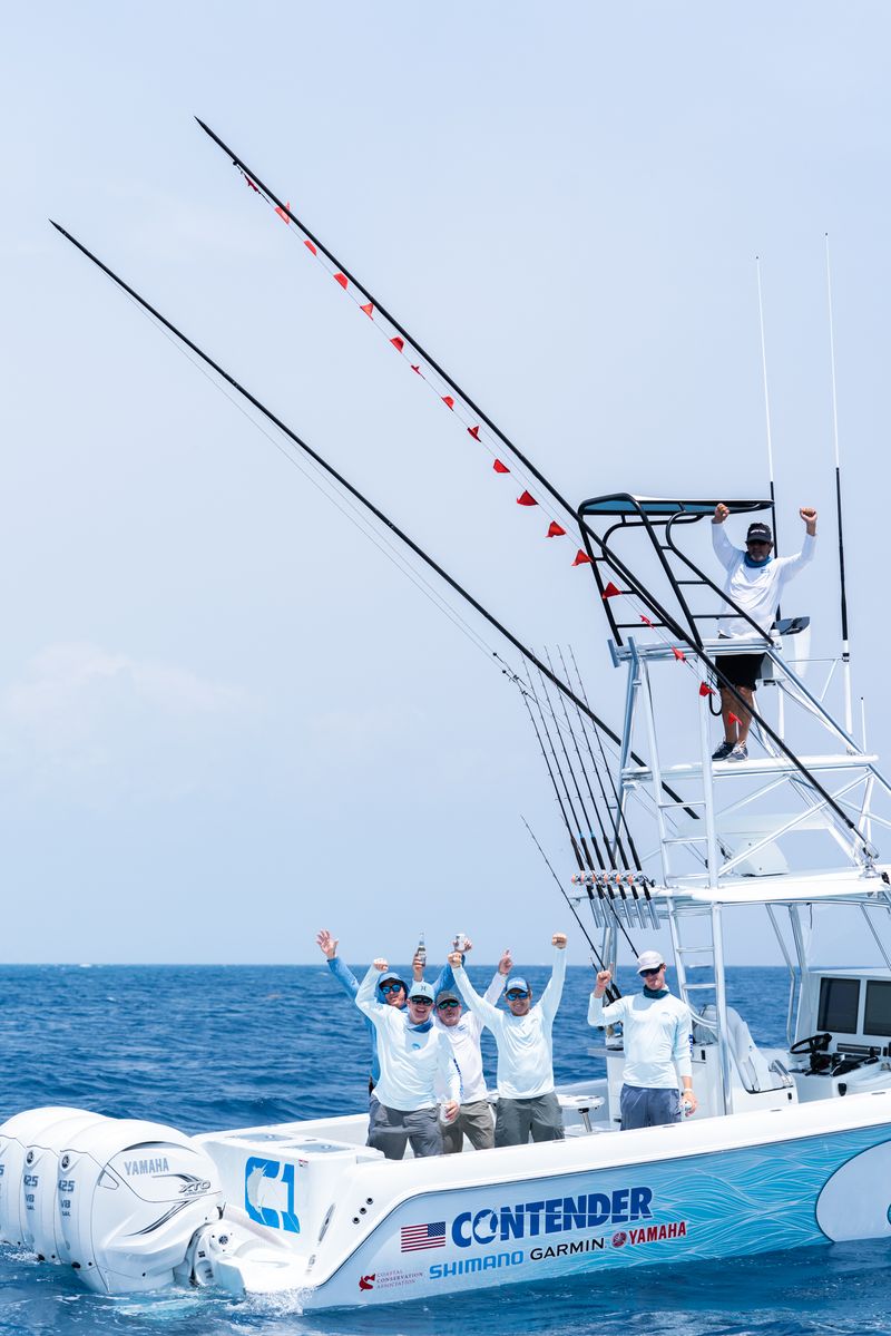 A group of people on a white sportfishing boat named "Contender" wave at the camera while out at sea. The boat is equipped with multiple fishing rods and Yamaha motors.