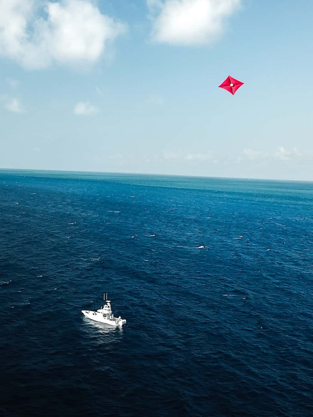 A white boat floats in the ocean under a blue sky with scattered clouds. A red diamond-shaped kite is flying above the water.