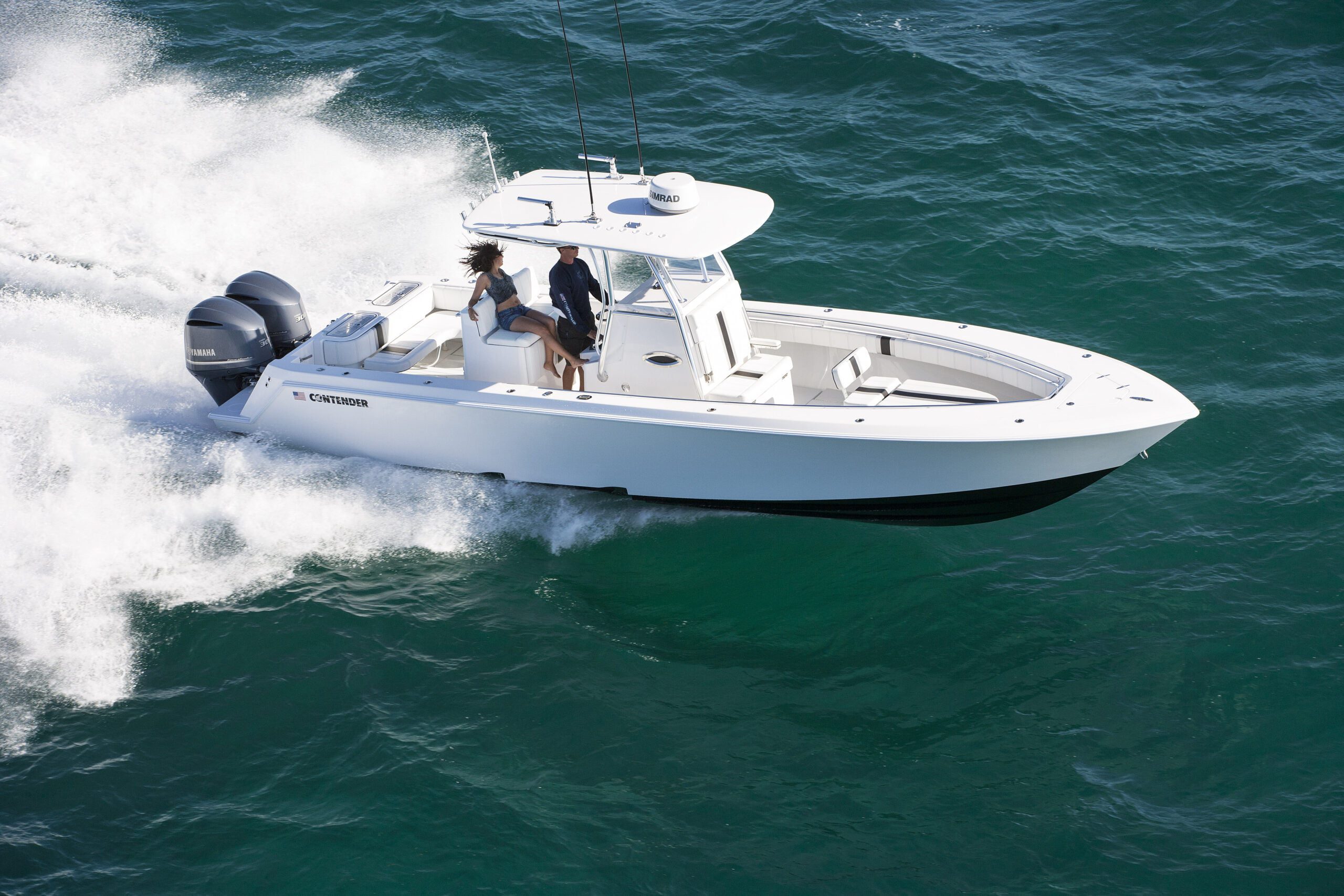 A white motorboat with two people onboard moves swiftly over the water, creating a wake behind it. The sea is calm and clear.