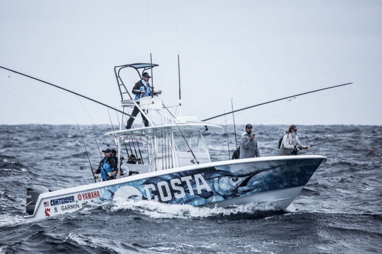 A fishing boat with several people onboard, equipped with fishing rods and branded with Costa, navigates through choppy waters.