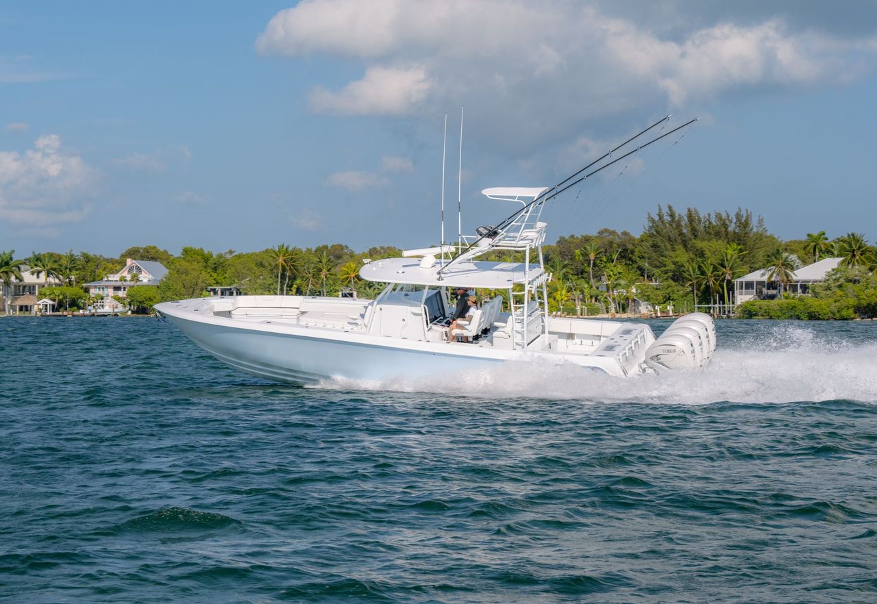 A white motorboat with multiple fishing rods cruises on blue water near a shoreline with houses and trees.