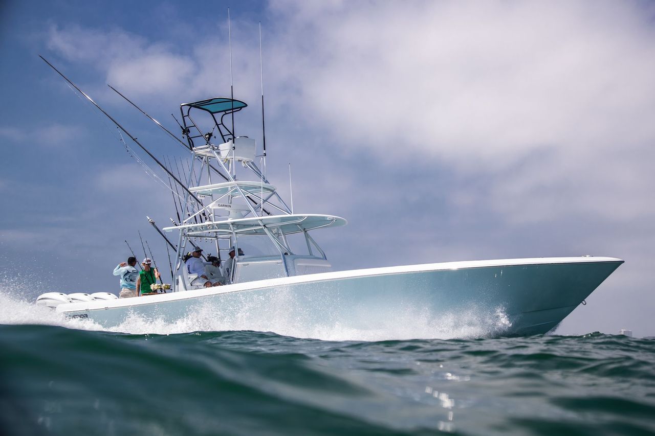 A white fishing boat with multiple fishing rods is navigating through the water under a partly cloudy sky. Several people are on board, some wearing hats and life jackets.