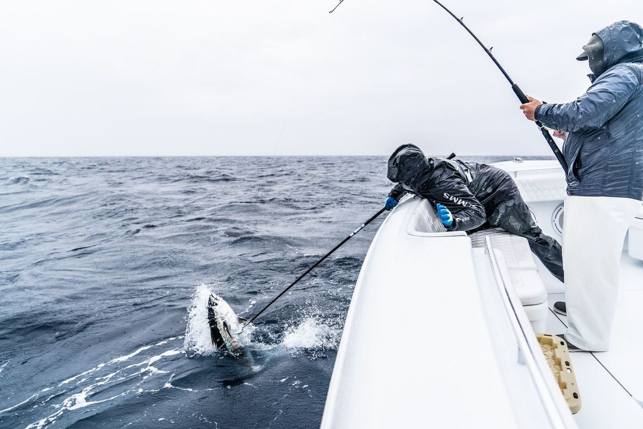 Two people fishing on a boat; one person holds a fishing rod while the other uses a net to capture a fish from the water. The scene takes place on a cloudy day with choppy waves.