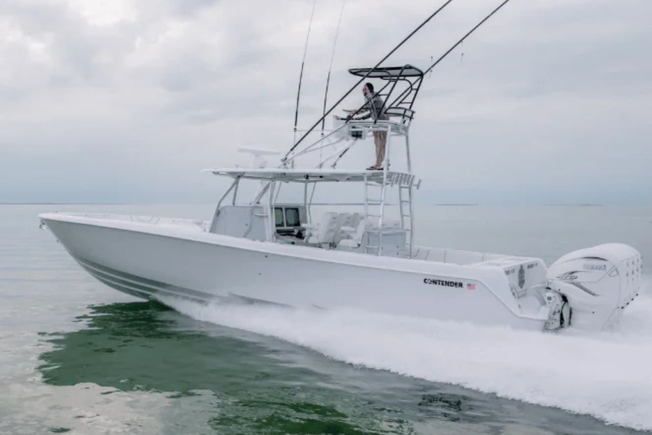 A white fishing boat with multiple fishing rods is moving swiftly across calm waters, with one person visible on an elevated platform piloting the vessel.