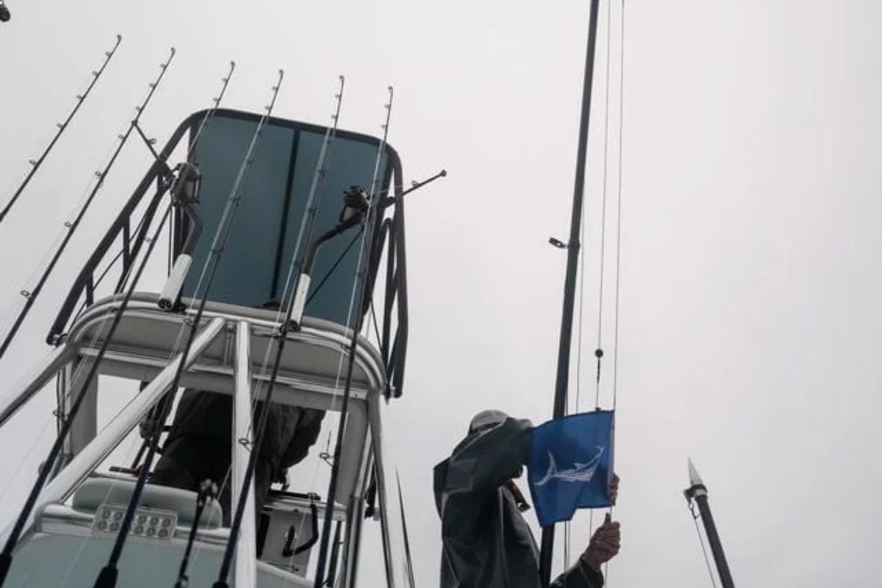 A person on a boat deck hoists a blue flag featuring a marlin graphic. Several fishing rods are visible, secured around the deck. The sky is overcast.