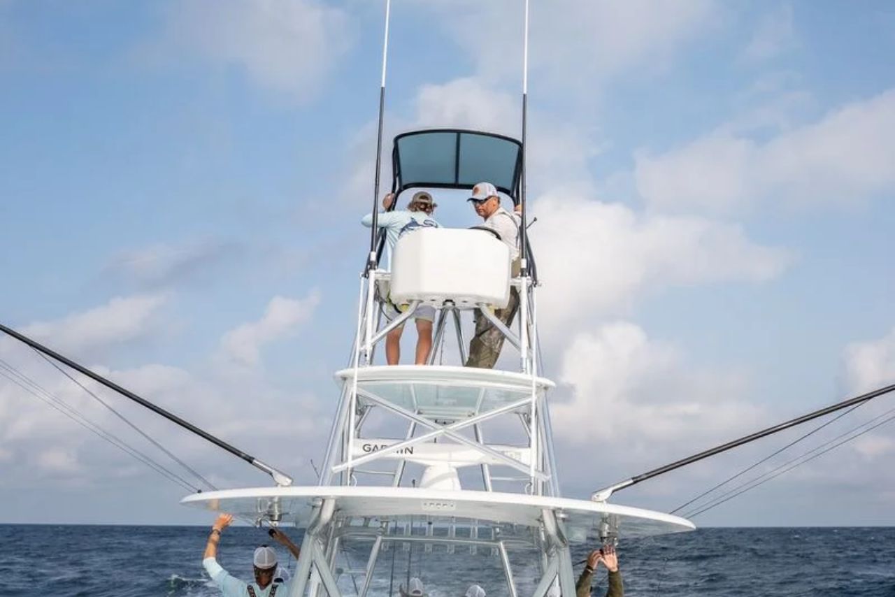 Two people stand at the top of a tall structure on a fishing boat, while two others work on the deck below, surrounded by the ocean under a partly cloudy sky.