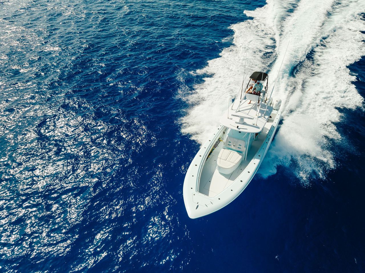 A white speedboat travels quickly across a deep blue ocean, leaving a frothy wake behind it. Two people are visible aboard the boat.