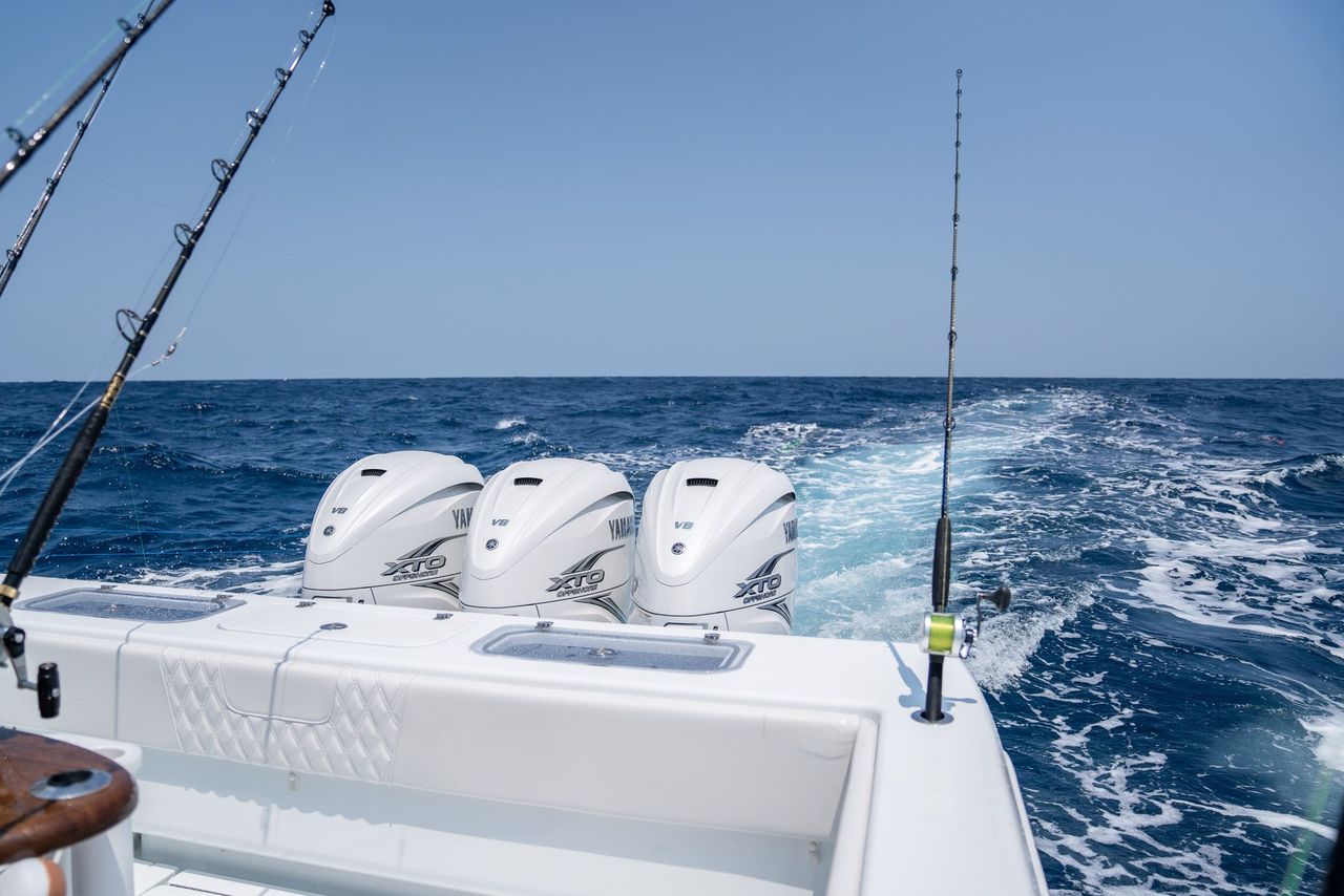 The rear view of a motorboat with three outboard engines and two fishing rods in a holder, moving through open water, leaving a wake.