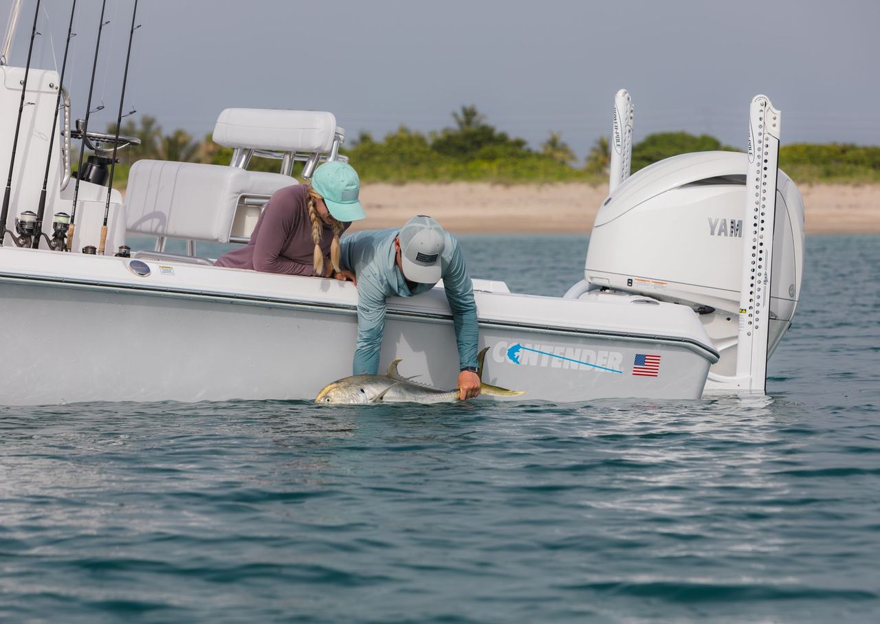Two people on a boat are holding a fish near the water's surface, with one person preparing to release it. The boat has fishing rods and a visible Yamaha engine.