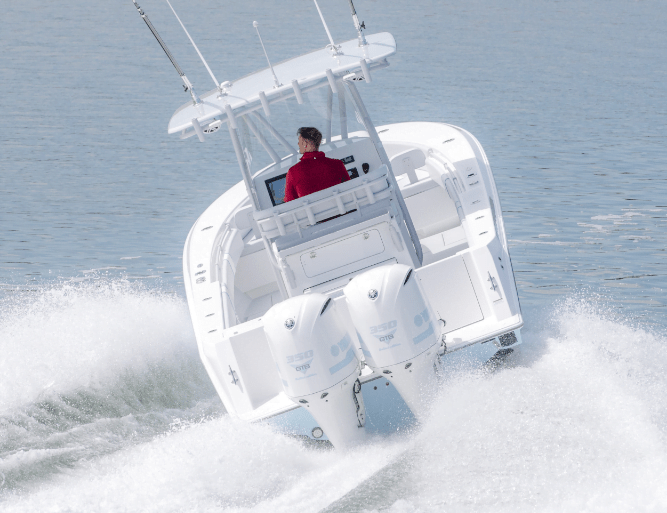 A person in a red jacket is driving a white motorboat with two engines, quickly moving on the water with wake visible behind.