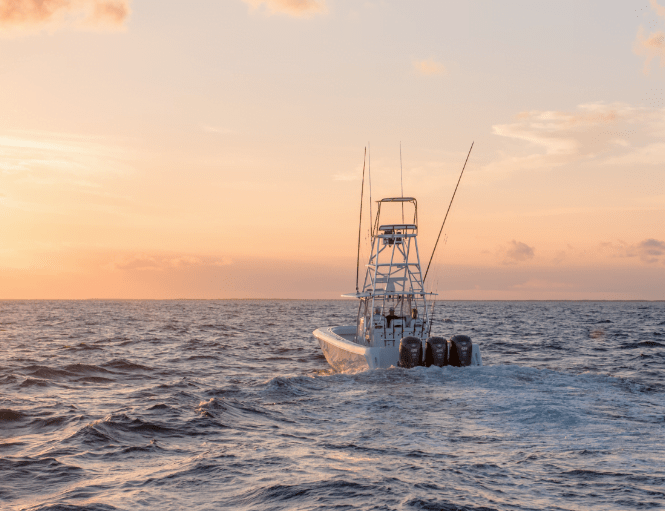 A motorboat equipped with fishing rods sails on the ocean during a sunset, with an orange and yellow sky in the background.