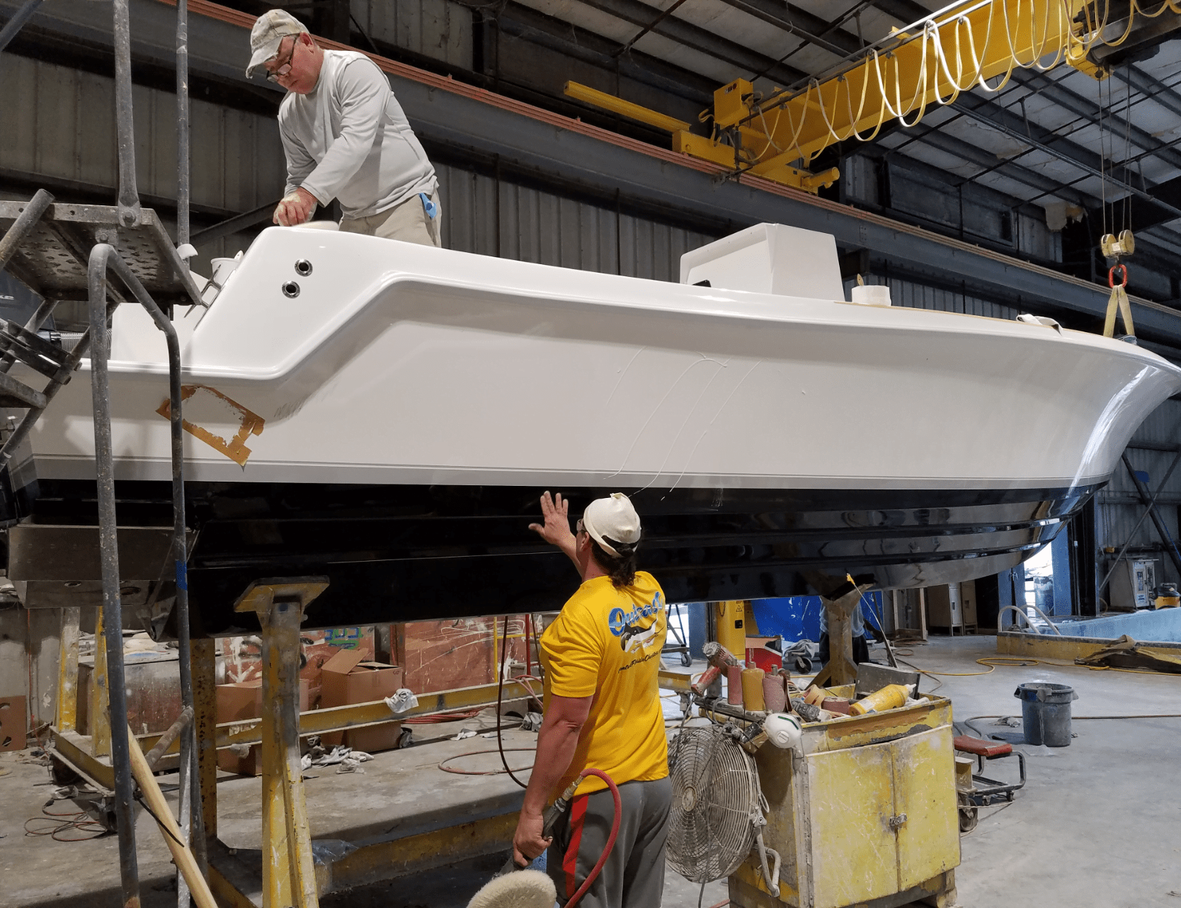 Two workers in a workshop are assembling and inspecting a white and black boat hull. One is on a scaffold while the other stands below, touching the hull. Various tools and equipment are visible around them.