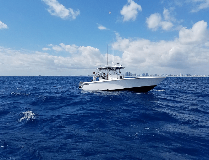 A white boat with a canopy floats on a vibrant blue ocean, with a distant city skyline visible on the horizon under a partly cloudy sky.