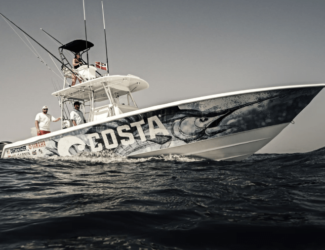 A fishing boat with "COSTA" and a large fish graphic on its side navigates through choppy waters, with two people on board.