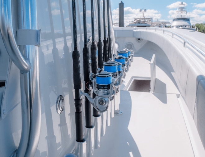 Several fishing rods with blue reels are neatly secured on a boat against a reflective glass wall, with other docked boats visible in the background under a partly cloudy sky.