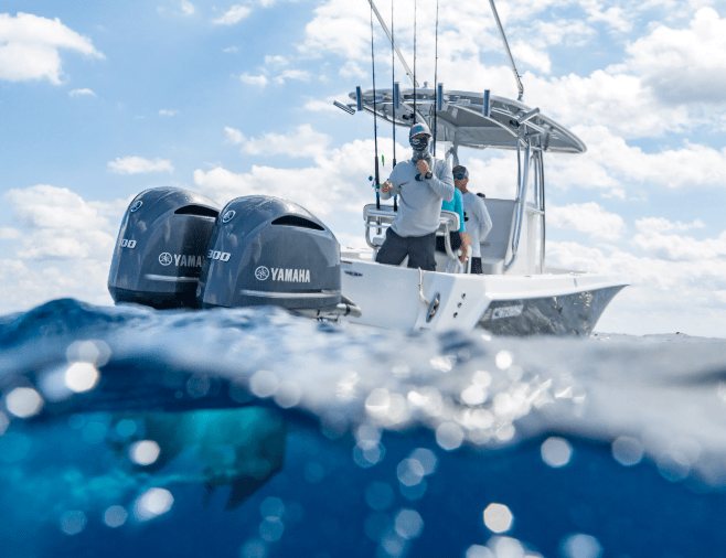 Two people stand on a boat equipped with Yamaha outboard motors, fishing in open water under a partly cloudy sky. The water partially covers the lower part of the image.