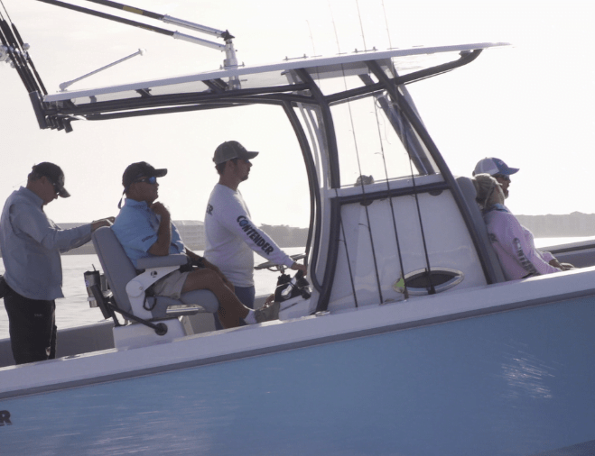 Four people are on a boat. Two are seated, and two are standing. The boat is on water under a light sky, with fishing rods visible.