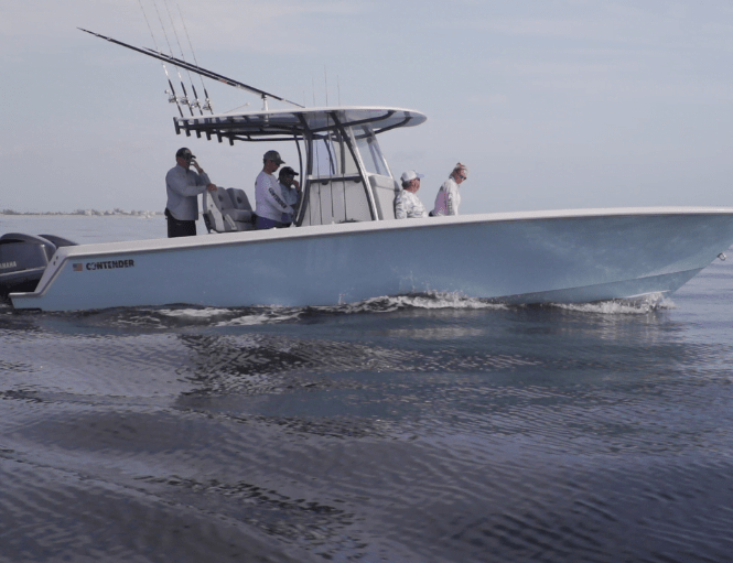 A group of people are on a motorized fishing boat with rods and equipment. The boat, labeled "Contender," is moving across calm water.