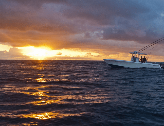 A small boat with fishing gear is seen on a calm ocean at sunset, with the sun partially obscured by clouds on the horizon.