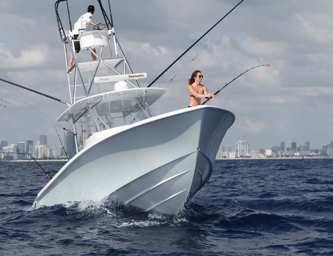 Two people fishing from a white boat in open water with a city skyline in the background. One is at the top of the boat’s tower, and the other is standing at the bow holding a fishing rod.