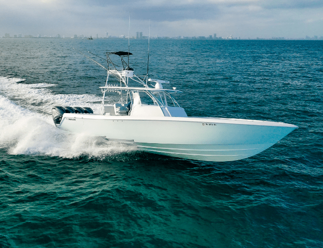 A white powerboat speeds through the ocean, with a city skyline visible in the distant background under a cloudy sky.
