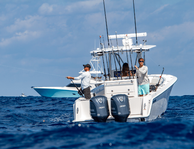 Two people fishing on a motorboat in open water with another boat visible in the background under a sunny sky.