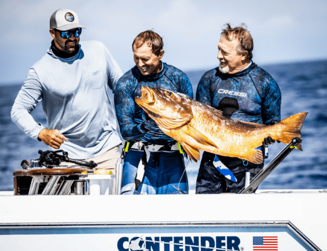 Three men standing on a boat hold a large fish, smiling. The boat has "CONTENDER" written on the side with an American flag sticker. The ocean is visible in the background.