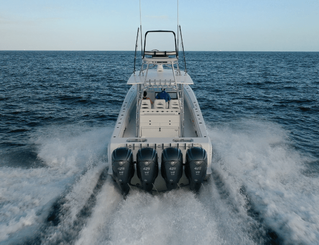 A large boat with four outboard motors speeds through open waters, leaving a wake behind. The boat has a central console and seating area, with two individuals seated.