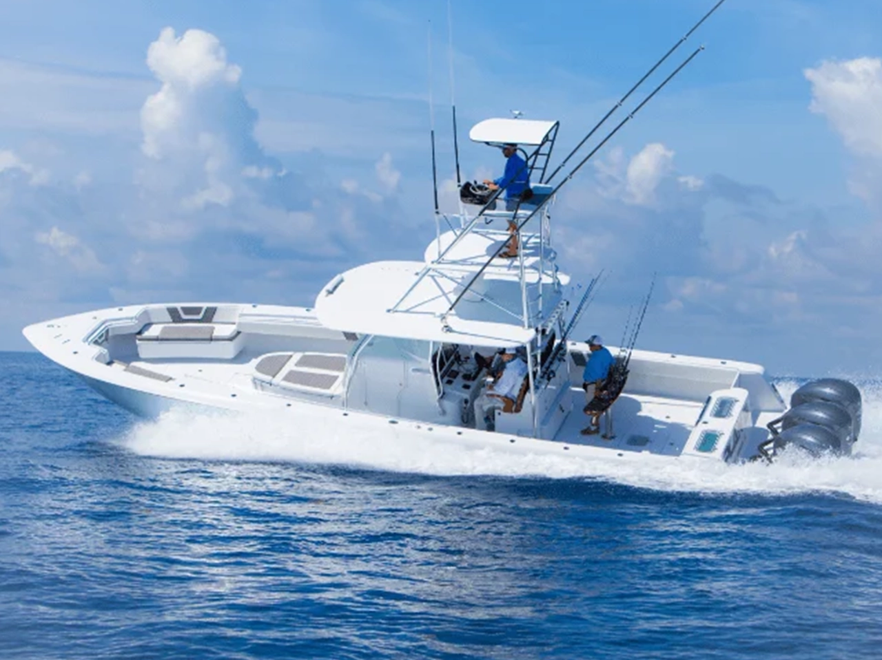 A white motorboat with multiple anglers, equipped with fishing rods and outboard engines, moves quickly across a calm blue ocean.
