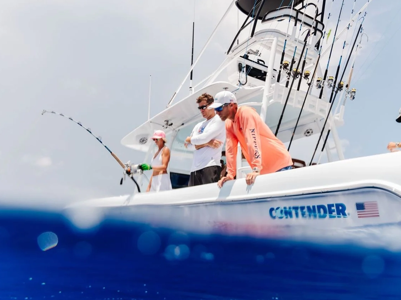 Four people on a boat are engaged in fishing, with one person actively reeling in a line while the others observe. The boat is branded with "CONTENDER" and has multiple fishing rods.