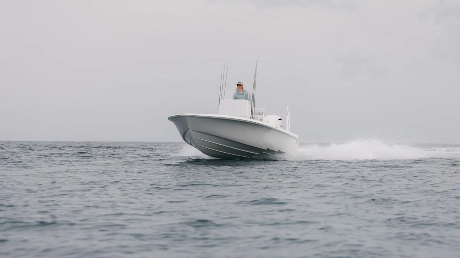 A person stands at the helm of a sleek 23BAY motorboat speeding across calm waters under a cloudy sky.